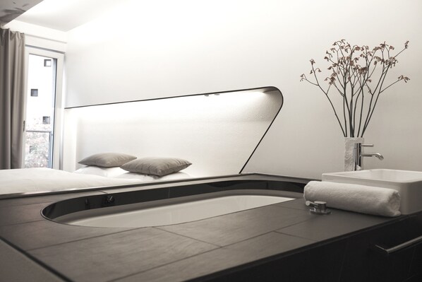 A modern hotel room with a curved bed, a built-in bathtub, a sink, and a vase with flowers.