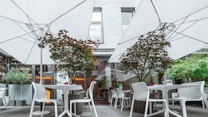 Un espace café en plein air avec des tables et des chaises blanches, entouré de murs de verre et abrité par des parasols blancs.