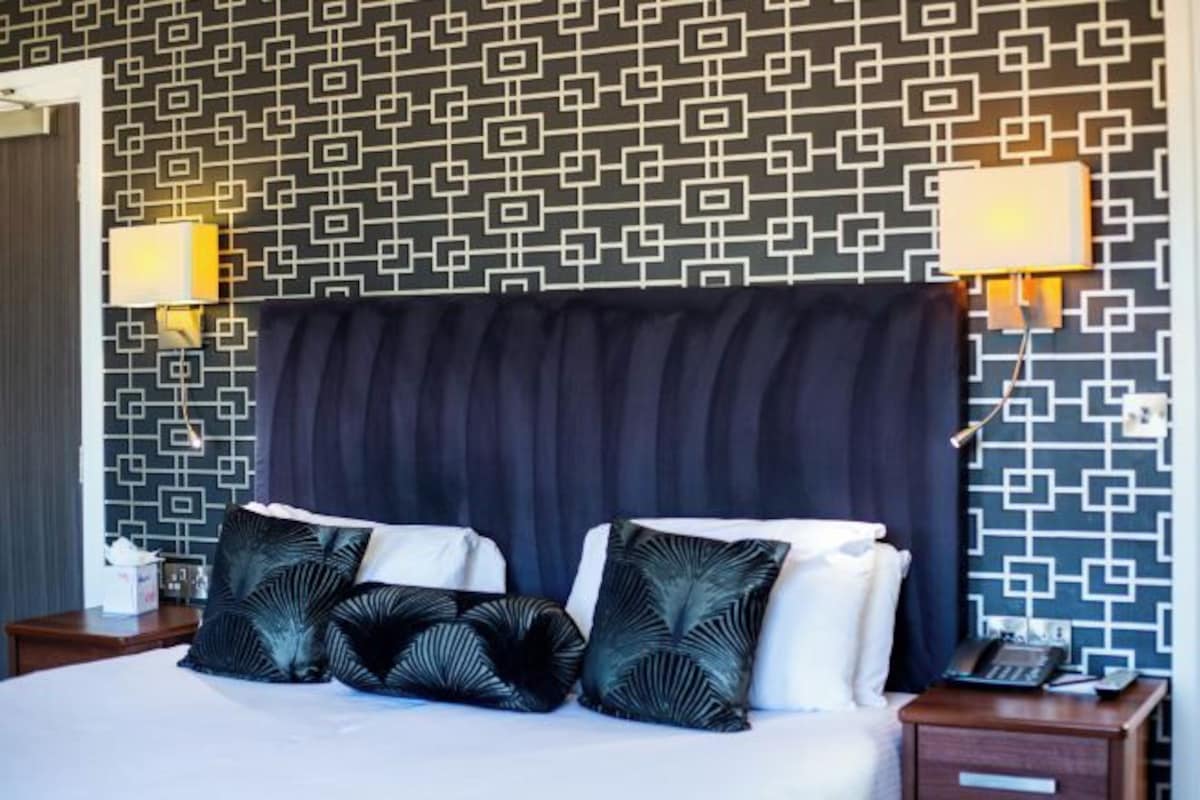 Una habitación de hotel con papel tapiz estampado, una cama grande con almohadas oscuras y blancas, dos lámparas de noche y una mesita de noche con un teléfono.
