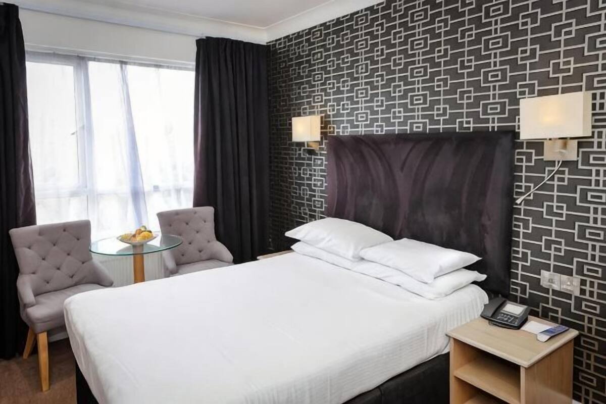 Camera d'albergo con un letto, due poltrone, un tavolino con della frutta, un comodino con una lampada e una parete decorata.
