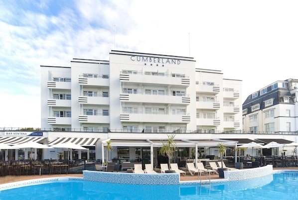Ein mehrstöckiges Hotel namens 'CUMBERLAND' mit Poolbereich, Außenbereich zum Sitzen und einer Bar namens 'BAR VENTANA'.