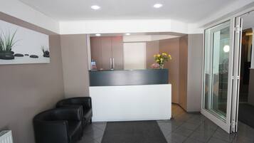 Un área de recepción con mostrador, dos sillones de cuero negro, alfombra gris y puerta de vidrio que da al exterior.
