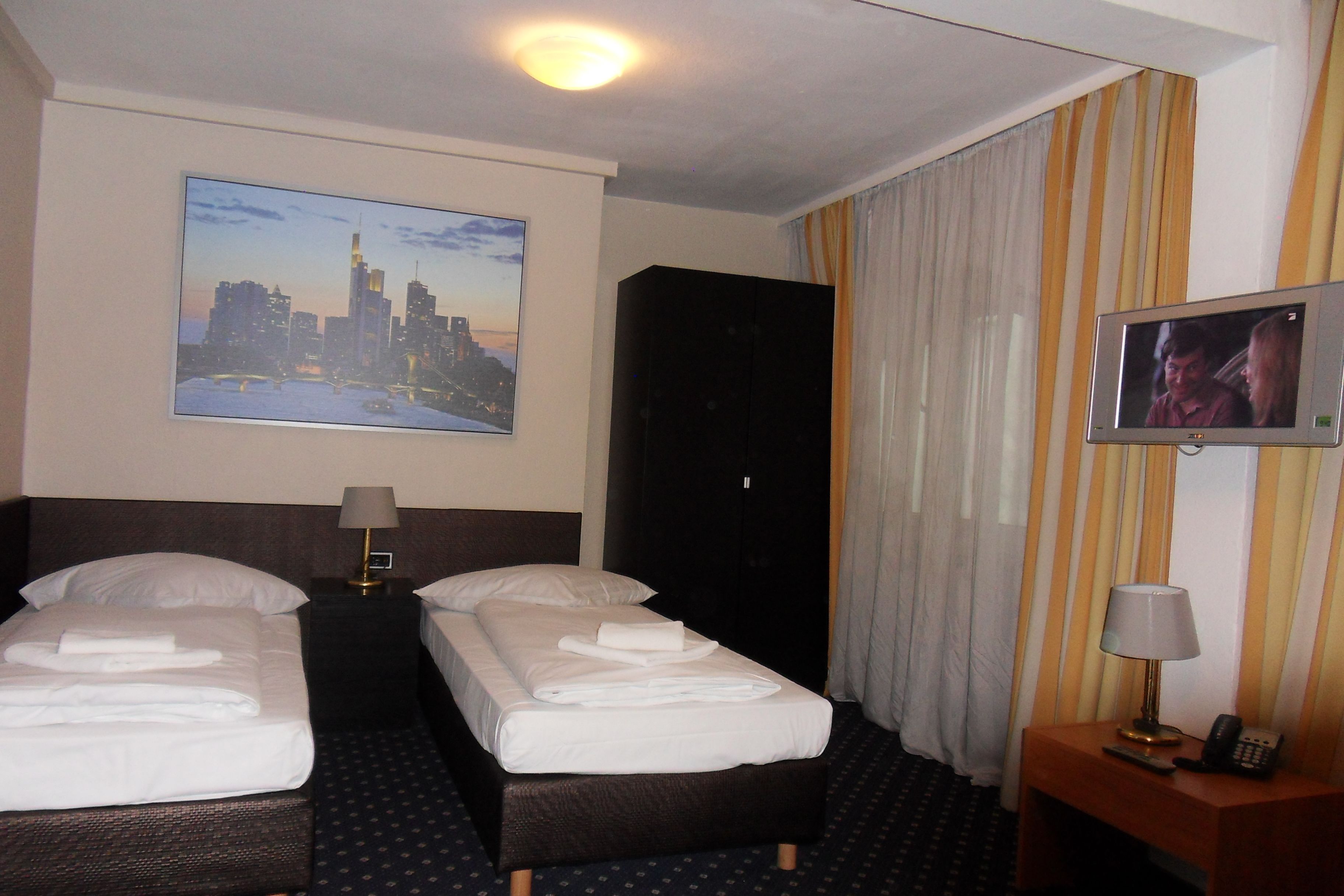 Habitación de hotel con dos camas, un cuadro de paisaje urbano, un televisor y un teléfono sobre una mesita de noche de madera.