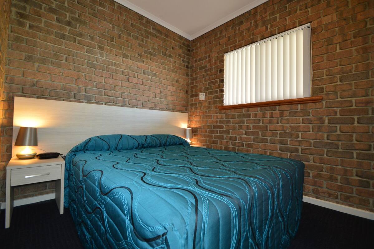 A bedroom with a brick wall, a bed with a blue quilt, a nightstand with a lamp, and a window with blinds.