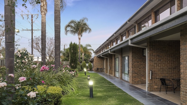 A building with a brick facade, outdoor seating area, and a well-maintained garden with flowers and palm trees. - Marion Motel and Apartments (Plympton Park)