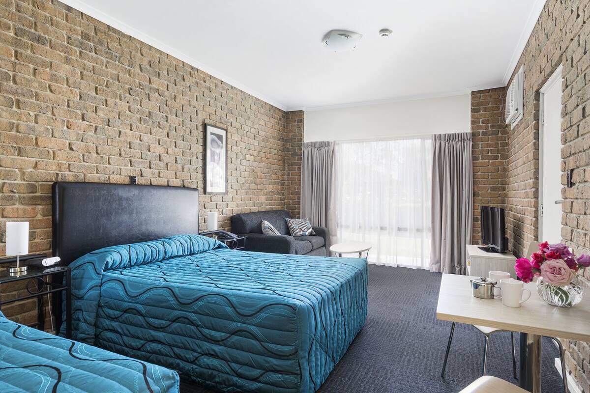 A hotel room with a brick wall, a bed with blue bedding, a sofa, a TV, a small table with a vase of flowers, and a window with curtains.