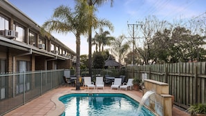 A swimming pool with a waterfall feature, surrounded by a fence and outdoor seating. - Marion Motel and Apartments (Plympton Park)
