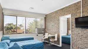 A modern room with a brick wall, a large window with blinds, a flat-screen TV mounted on the wall, a small table with chairs, and a sofa with blue bedding. - Marion Motel and Apartments (Plympton Park)