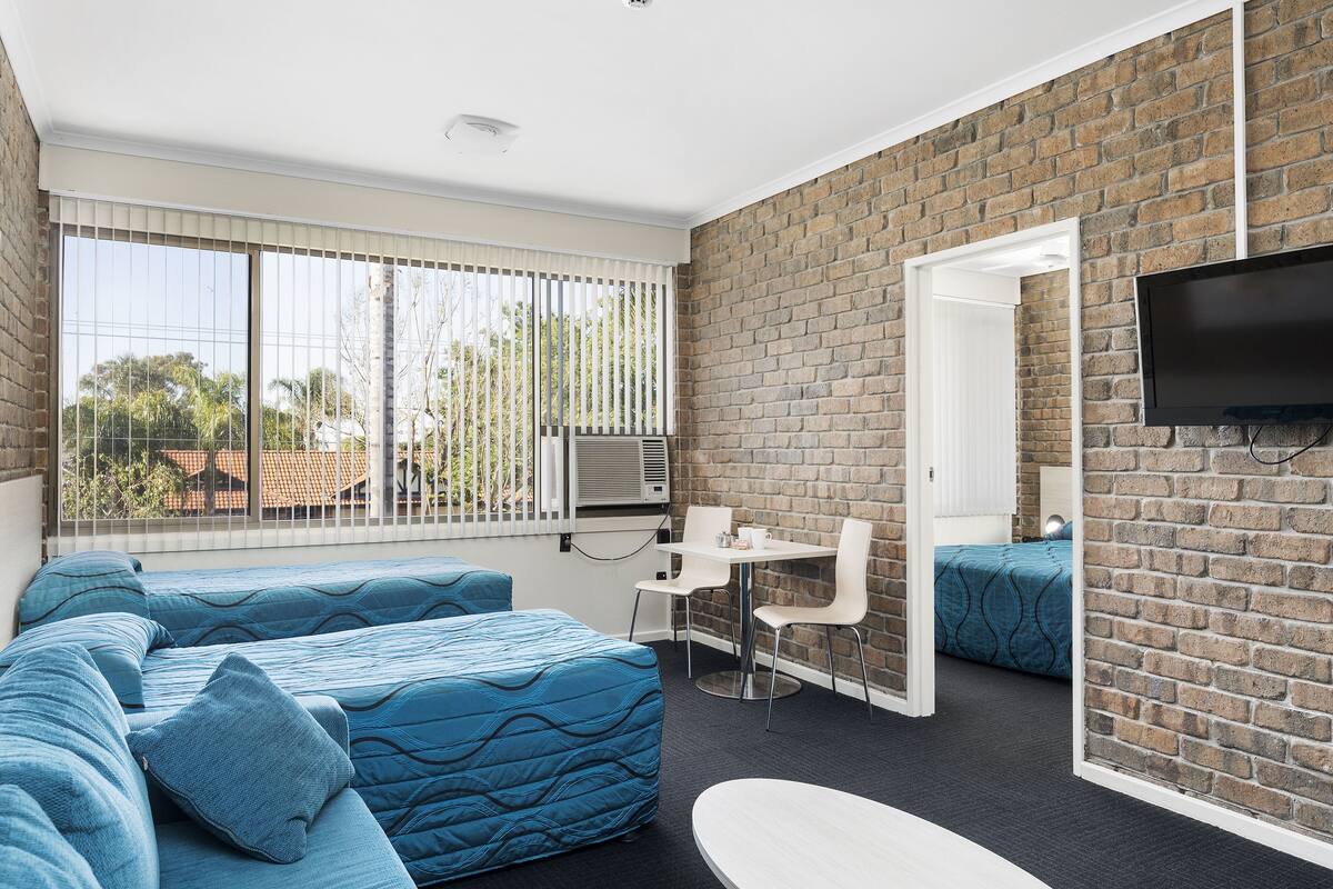 A modern room with a brick wall, a large window with blinds, a flat-screen TV mounted on the wall, a small table with chairs, and a sofa with blue bedding.