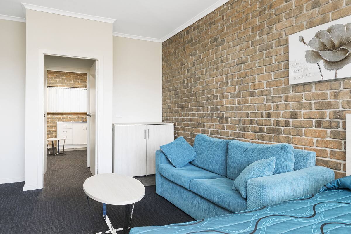 A modern living room with a brick wall, a blue sofa, a round table, and a kitchen area visible through an open doorway.