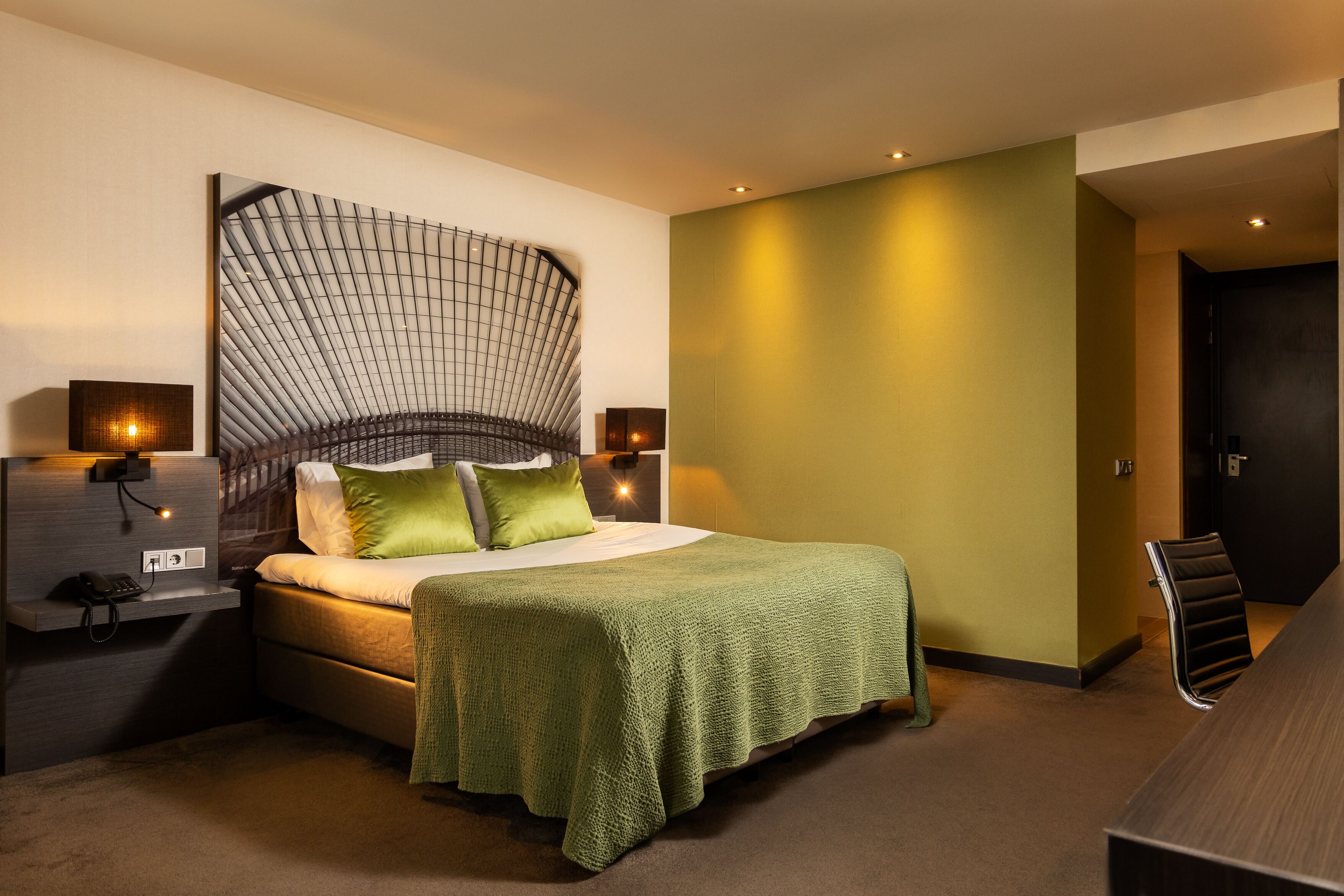 Una habitación de hotel moderna con un cabecero curvo, una manta verde, una mesita de noche con una lámpara y una silla.