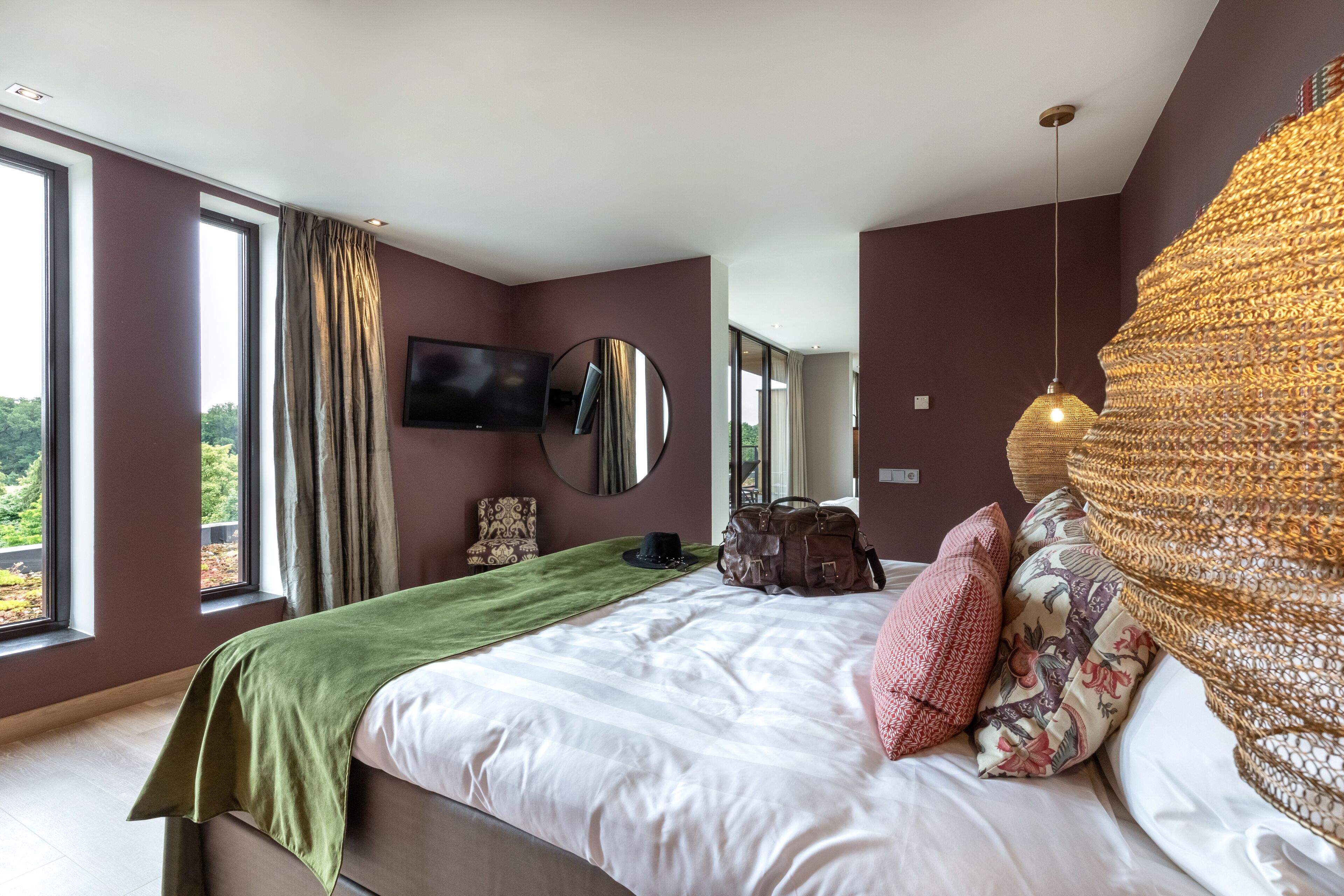 Ein modernes Schlafzimmer mit einem großen Bett, einem Flachbildfernseher, einem Spiegel und Blick auf Grünflächen.