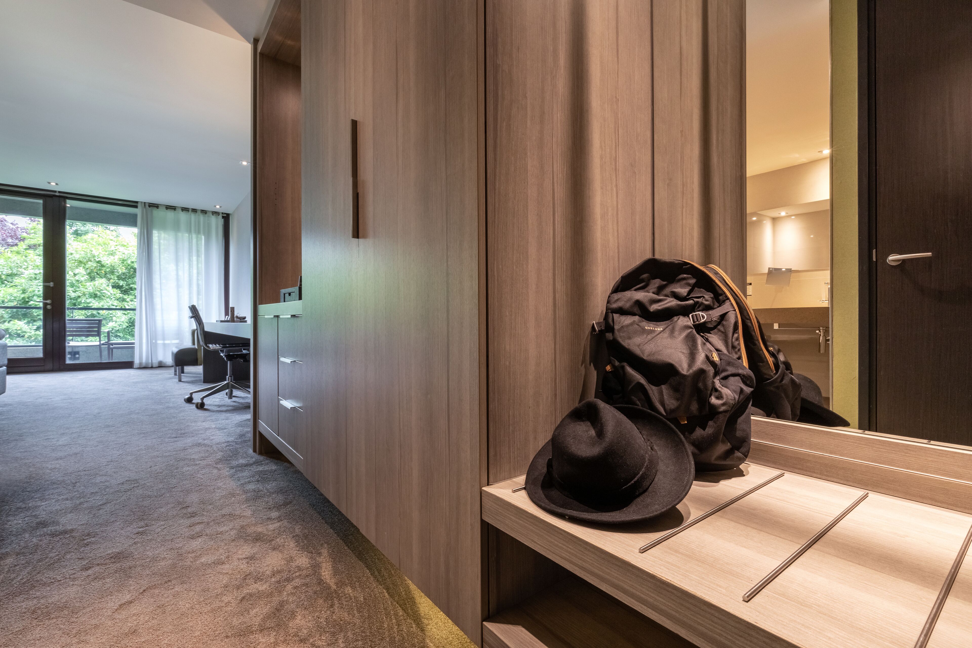 Ein Hotelzimmer mit Holzschrank, Schreibtisch und Stuhl sowie einem großen Fenster mit Vorhängen.