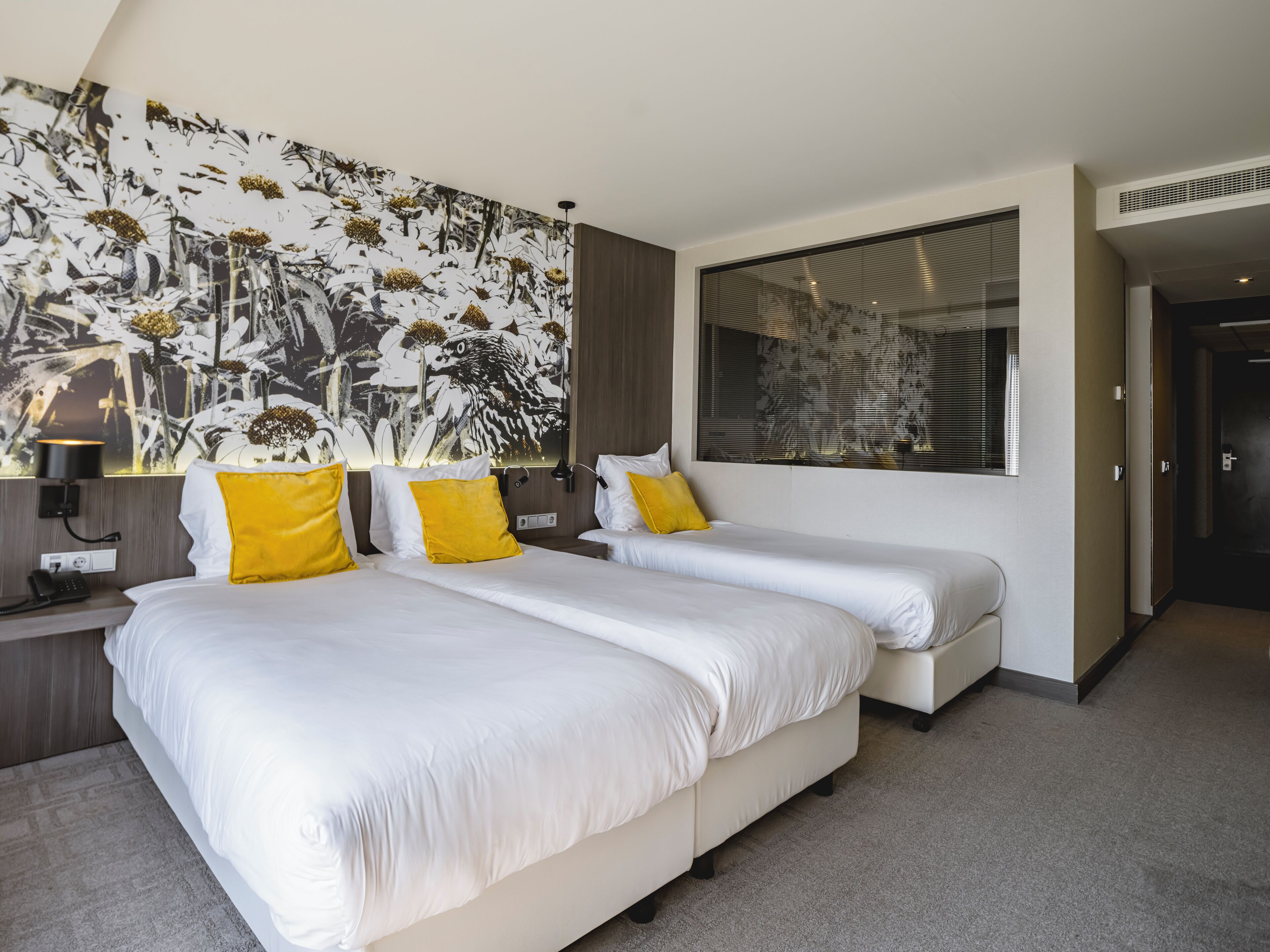 Une chambre d’hôtel avec deux lits, un grand décor mural floral, une fenêtre avec des stores et un design moderne.