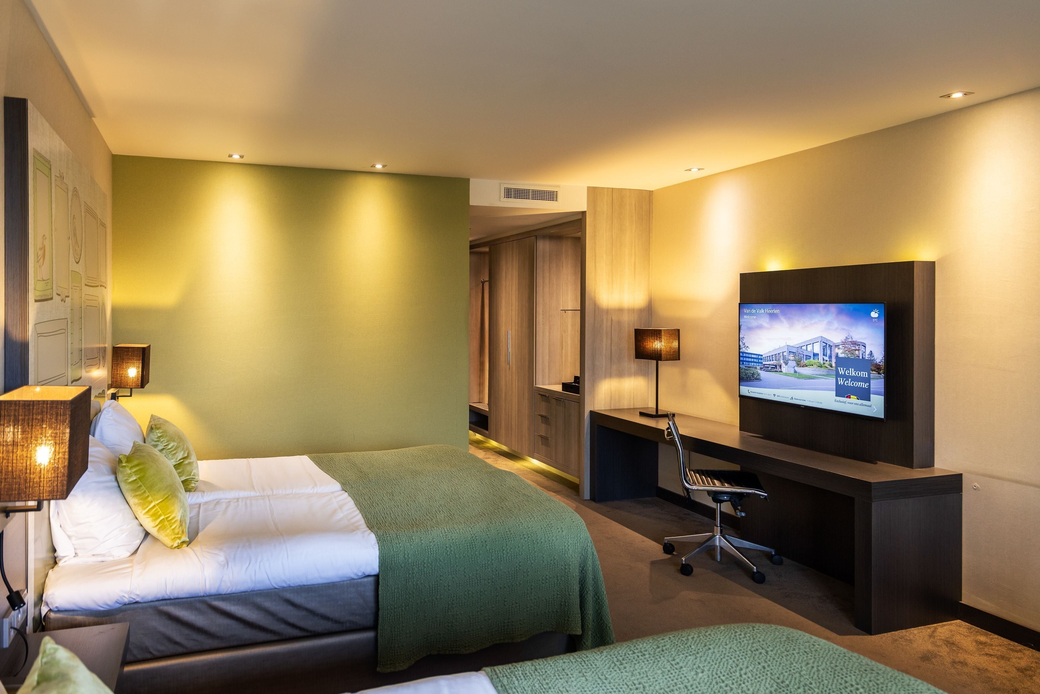 Ein Hotelzimmer mit zwei Betten, einem Schreibtisch, einem Fernseher und einem Wandfernseher.