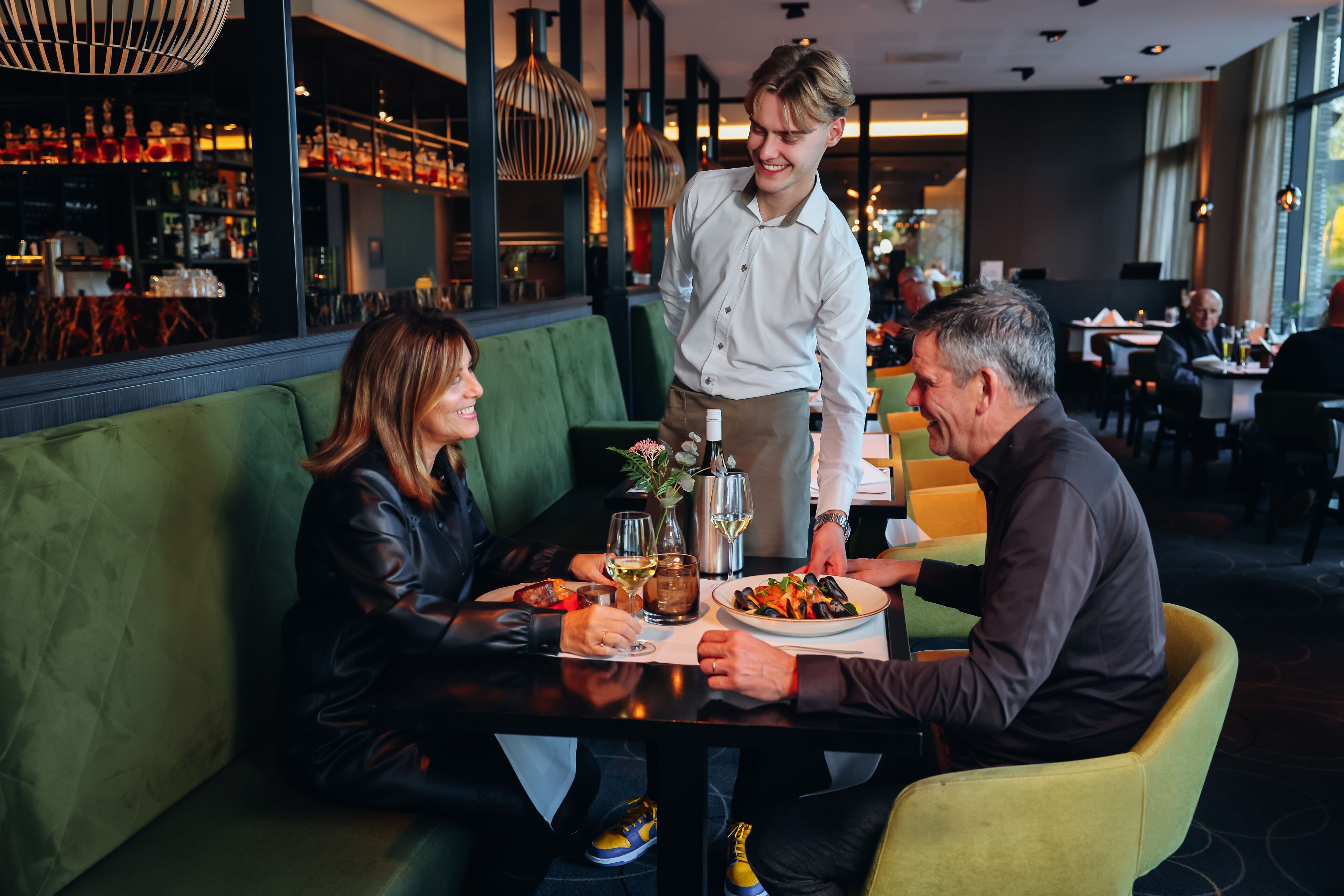 Um restaurante com assentos em cabines verdes, um bar ao fundo e um garçom servindo uma refeição.