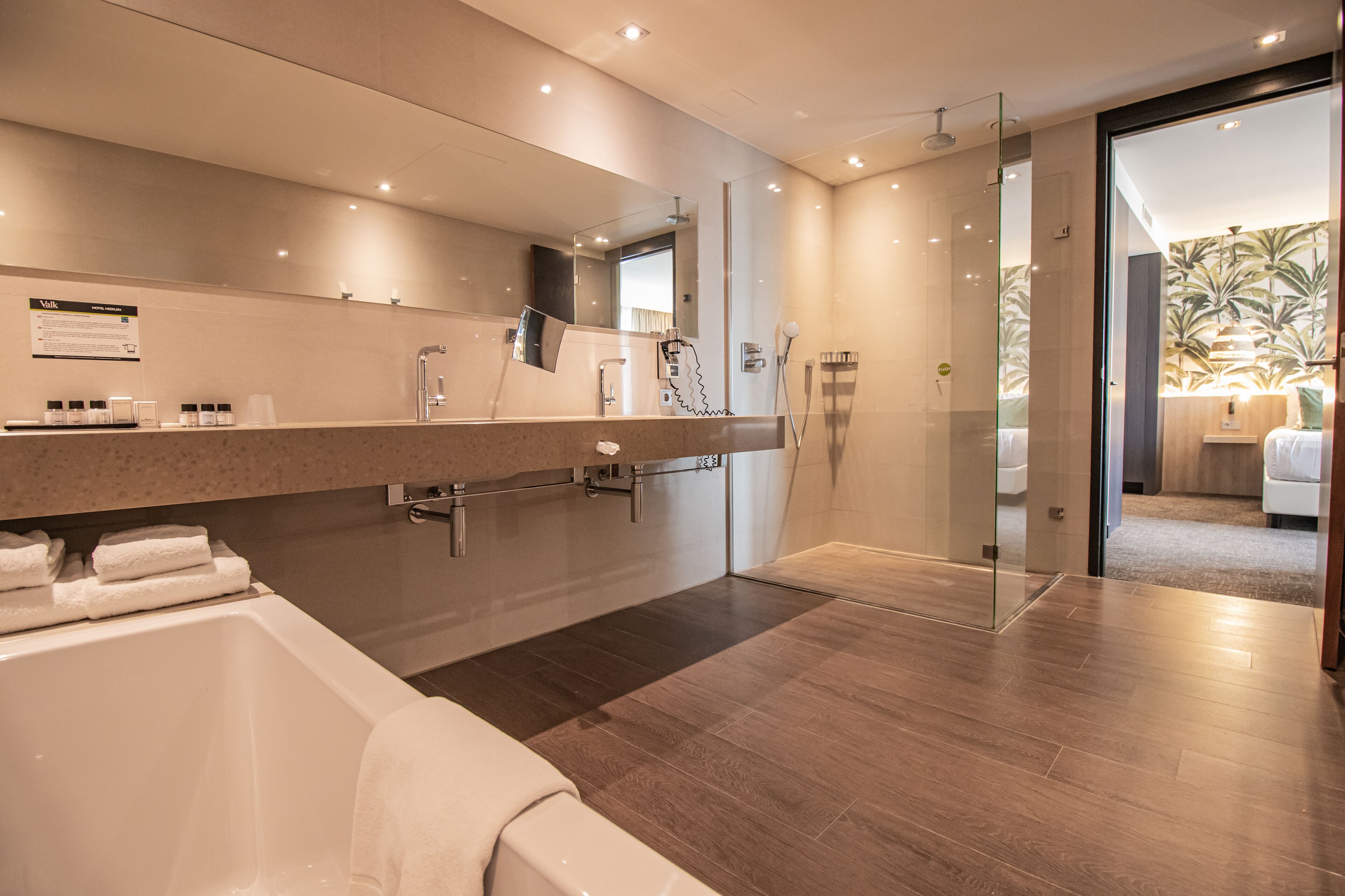 A modern bathroom with a bathtub, a large mirror, a glass shower enclosure, and a view of a bedroom with a bed and a plant.