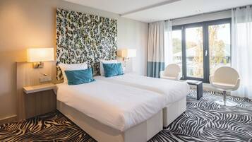 A modern hotel room with a large bed, two bedside tables with lamps, a zebra-patterned carpet, and a view of the outdoors through large windows.
