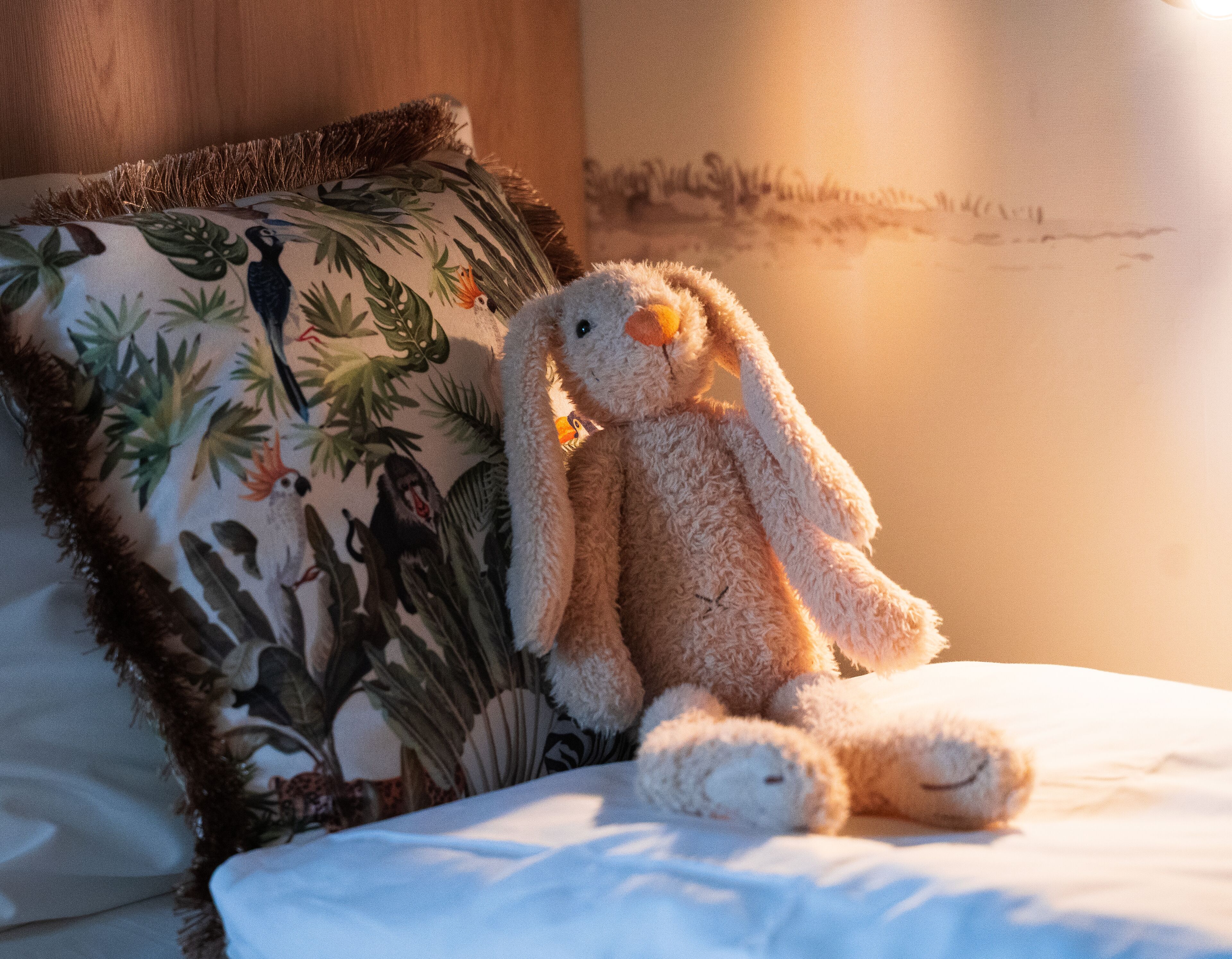Un peluche sobre una cama con un cojín floral y una pared con un mural.