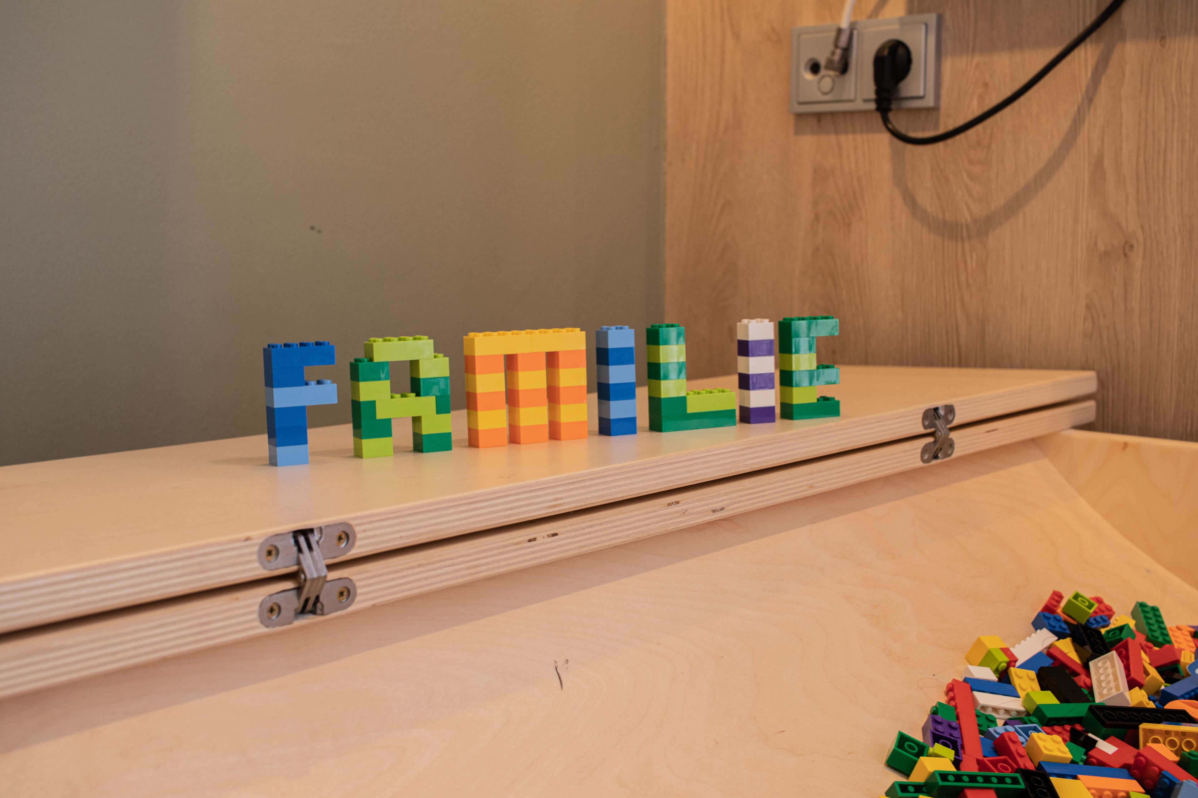 Een houten plank waarop LEGO letters het woord “FAMILY” spellen, en een stapel LEGO stukjes op de vloer.