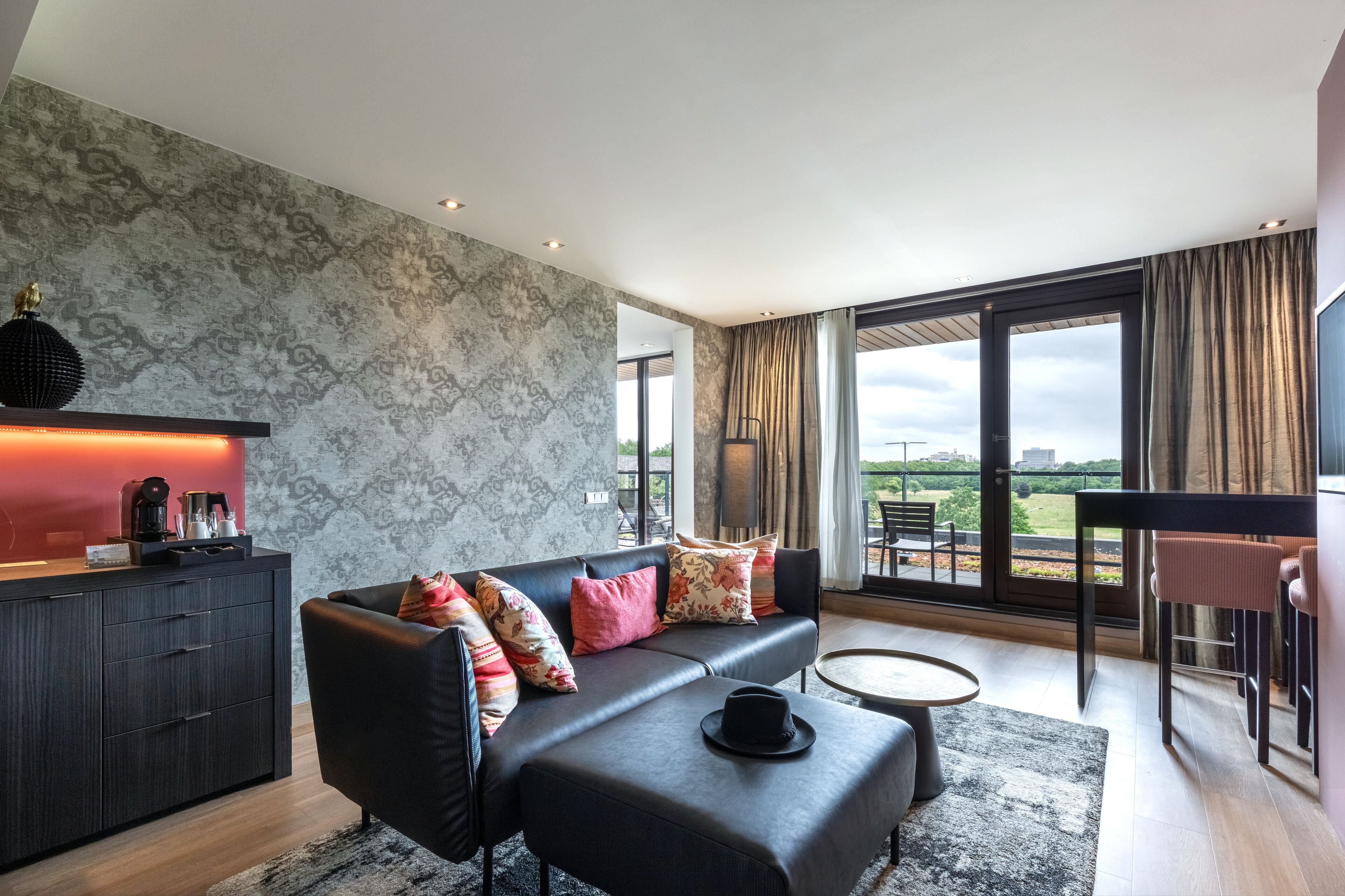 A modern living room with a sofa, ottoman, and coffee table. There is a view of the outdoors through large windows.