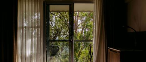 Une chambre à coucher avec un lit, une commode en bois et une porte-fenêtre coulissante donnant sur un balcon bordé d’arbres.