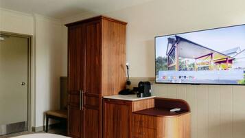 A hotel room with a wooden wardrobe, a small desk with a coffee maker, a television mounted on the wall, and a trash bin.