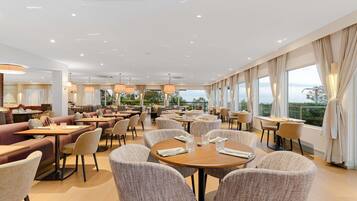 A spacious hotel dining area with round tables and cushioned chairs, large windows with curtains, and a view of the outdoors.