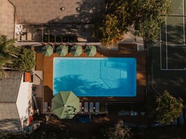 Vista aérea de una piscina rectangular con sillas de descanso y sombrillas, rodeada de árboles y un edificio.