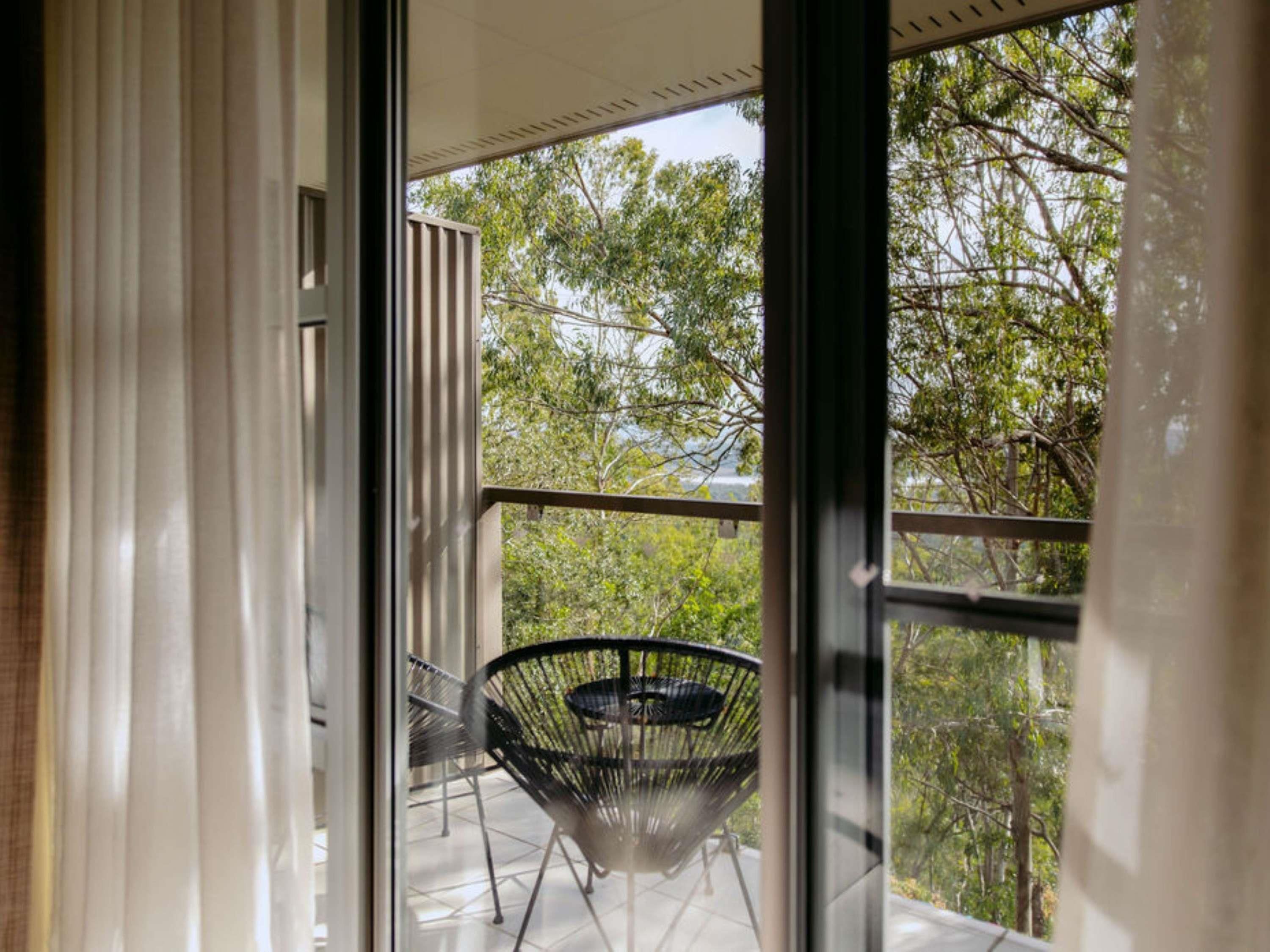 Vista a través de un ventanal hacia un balcón exterior con una silla de metal y una mesa.