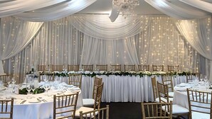Un salón de banquetes con mesas redondas, cortinas blancas y un candelabro.
