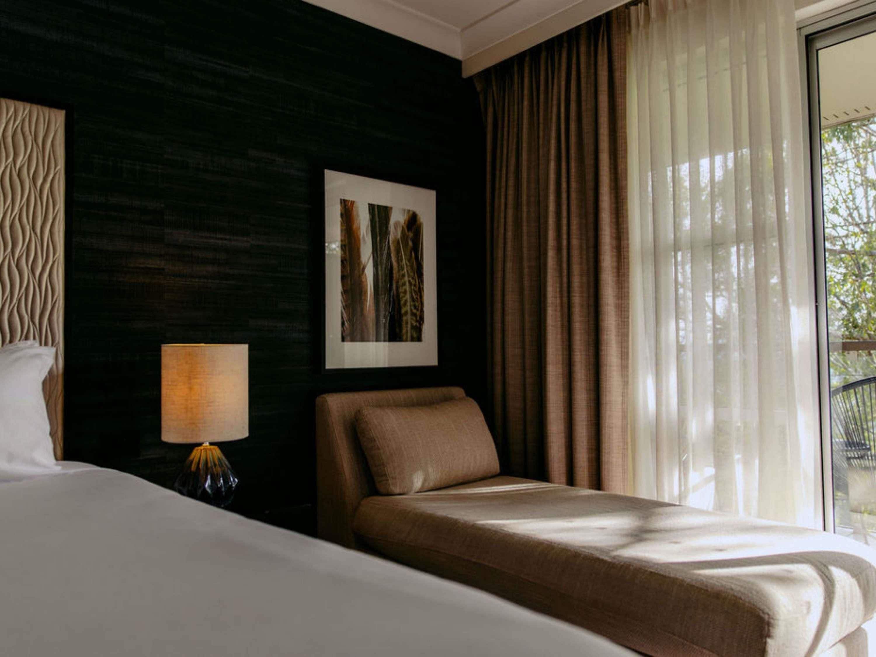 A hotel room with a bed, a bedside lamp, a chair, a framed picture on the wall, and a window with curtains.