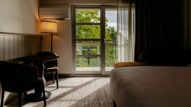 Habitación de hotel con cama, escritorio, silla, lámpara y puerta corrediza de vidrio que da a un área exterior.