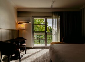 Habitación de hotel con cama, escritorio, silla, lámpara y puerta corrediza de vidrio que da a un área exterior.