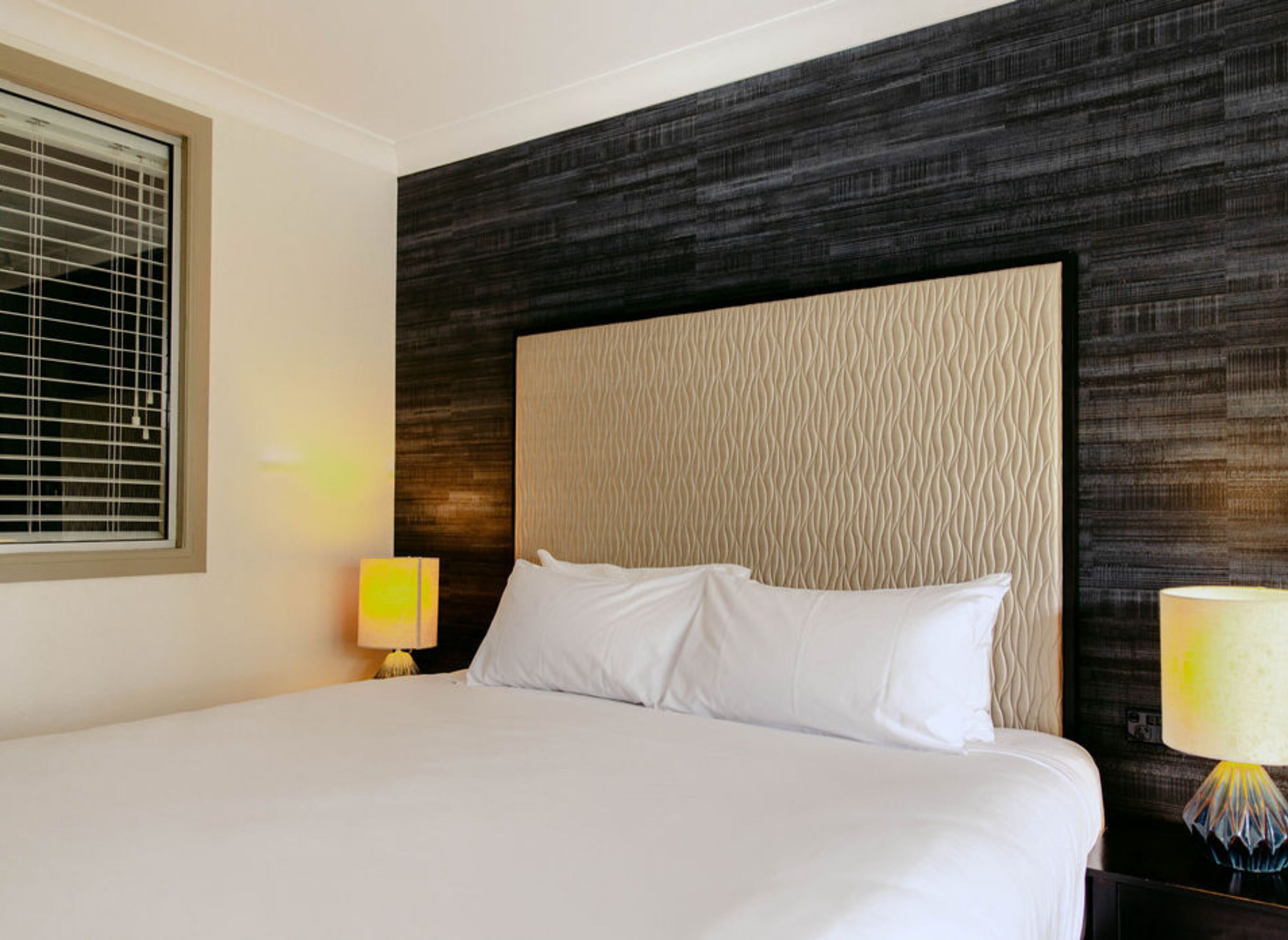 A hotel room with a large bed, a headboard with a textured pattern, a window with blinds, and two bedside lamps.