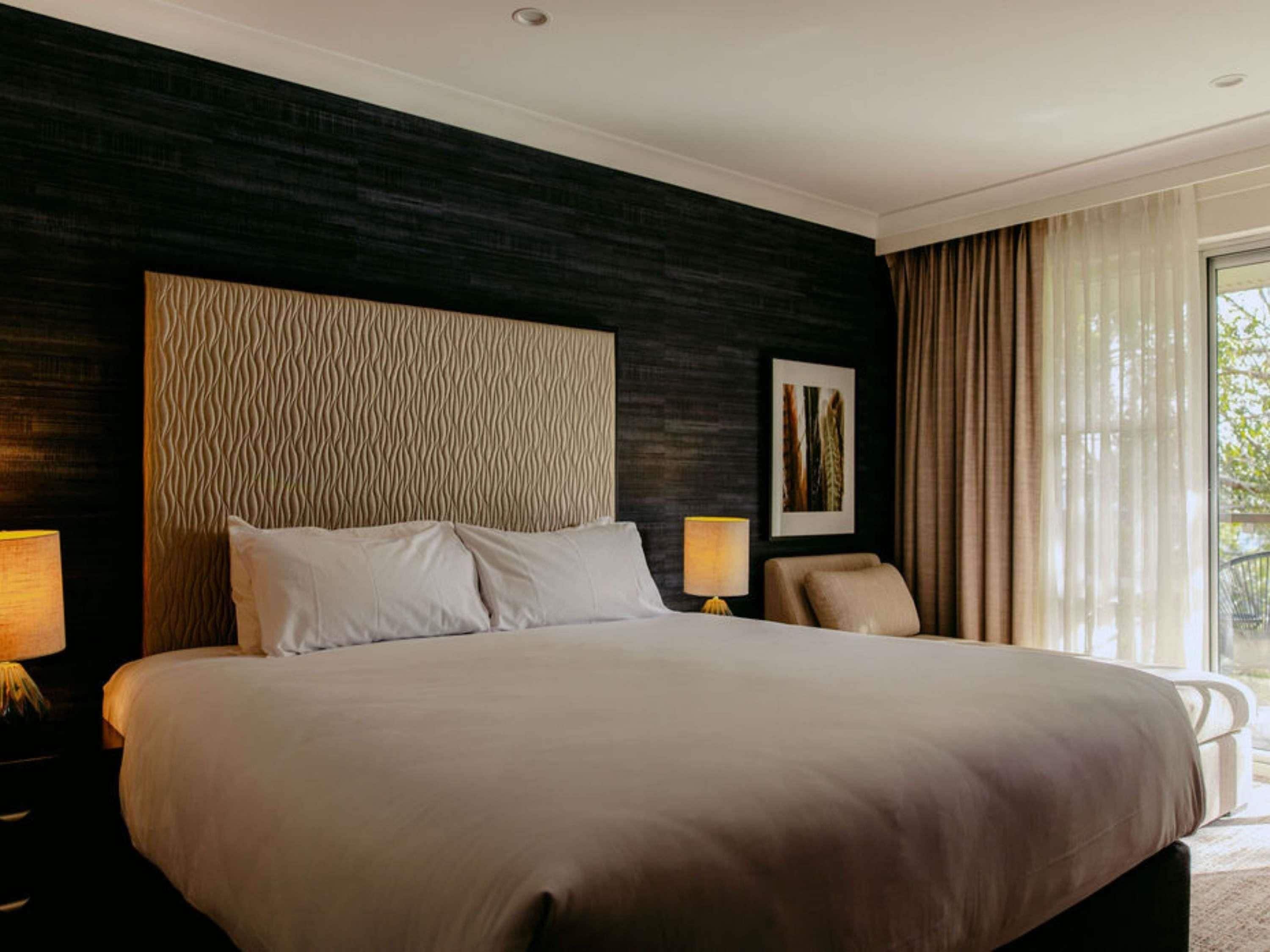 Habitación de hotel con una cama grande, dos lámparas de noche, una silla, una ventana con cortinas y una pared con textura.