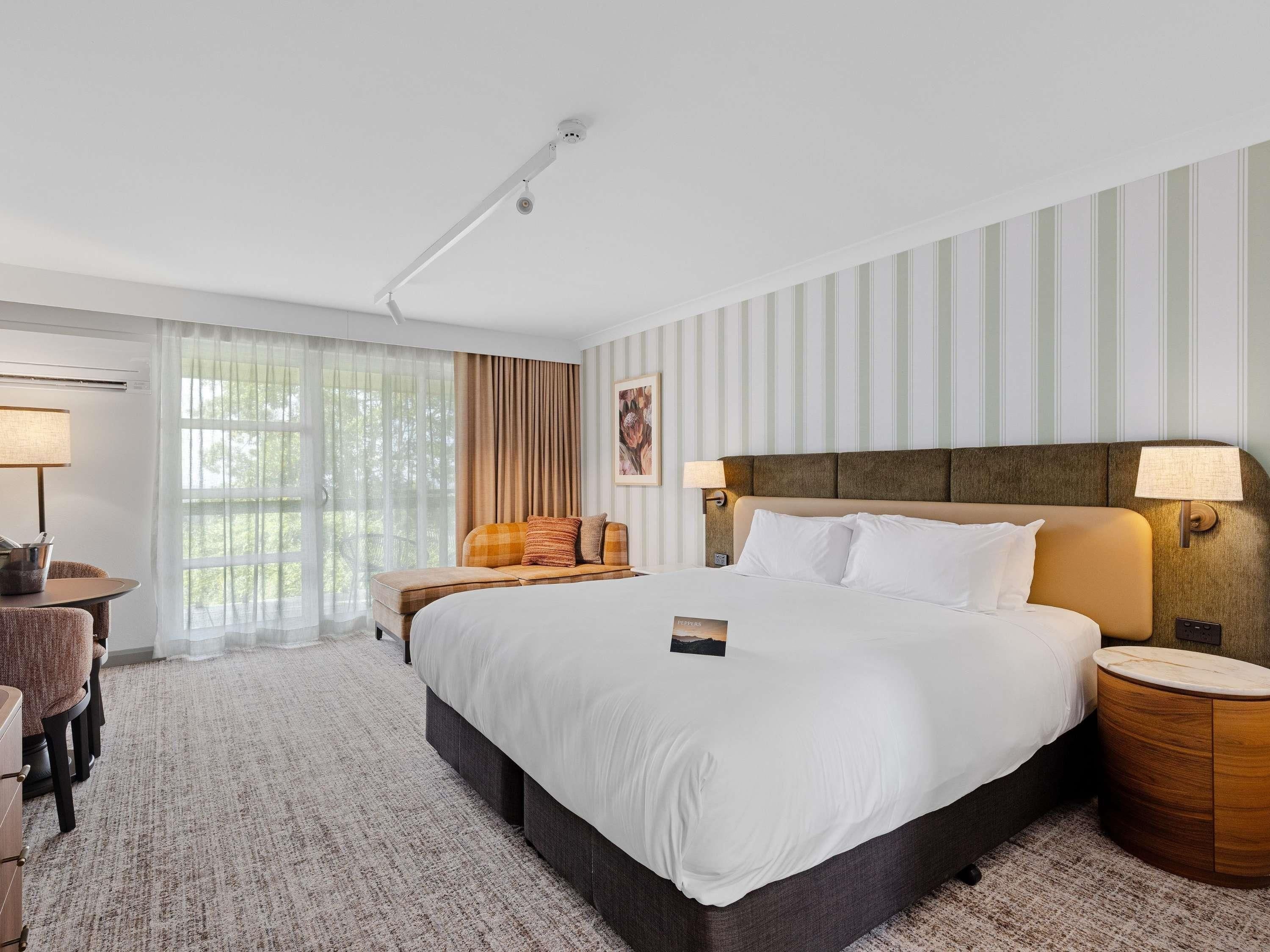 Una habitación de hotel con una cama grande, una mesita de noche, una silla, una lámpara y una ventana con cortinas.