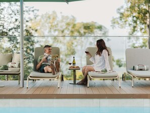 Dos personas sentadas en una terraza con piscina al frente, leyendo revistas y sosteniendo una copa de vino.