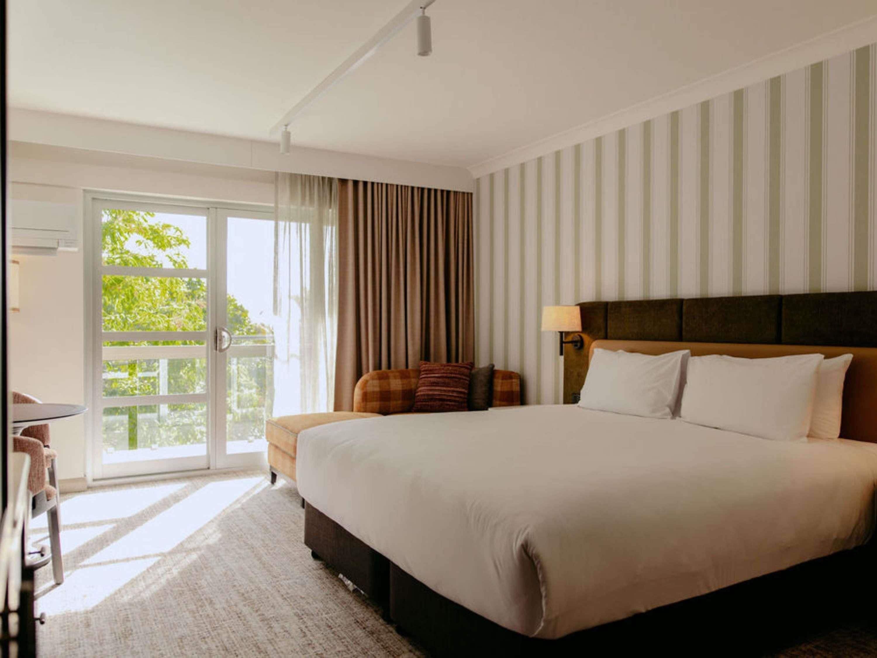 A hotel room with a large bed, a desk, and a view of greenery through the window.