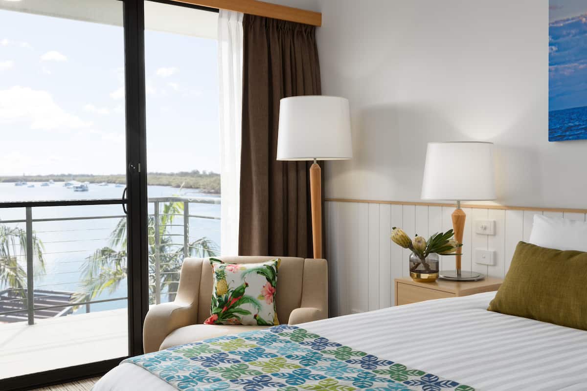 Une chambre d’hôtel avec un grand lit, deux lampes de chevet, une chaise avec un coussin à motifs floraux, une table de chevet avec un vase, et une vue sur la mer depuis un balcon.