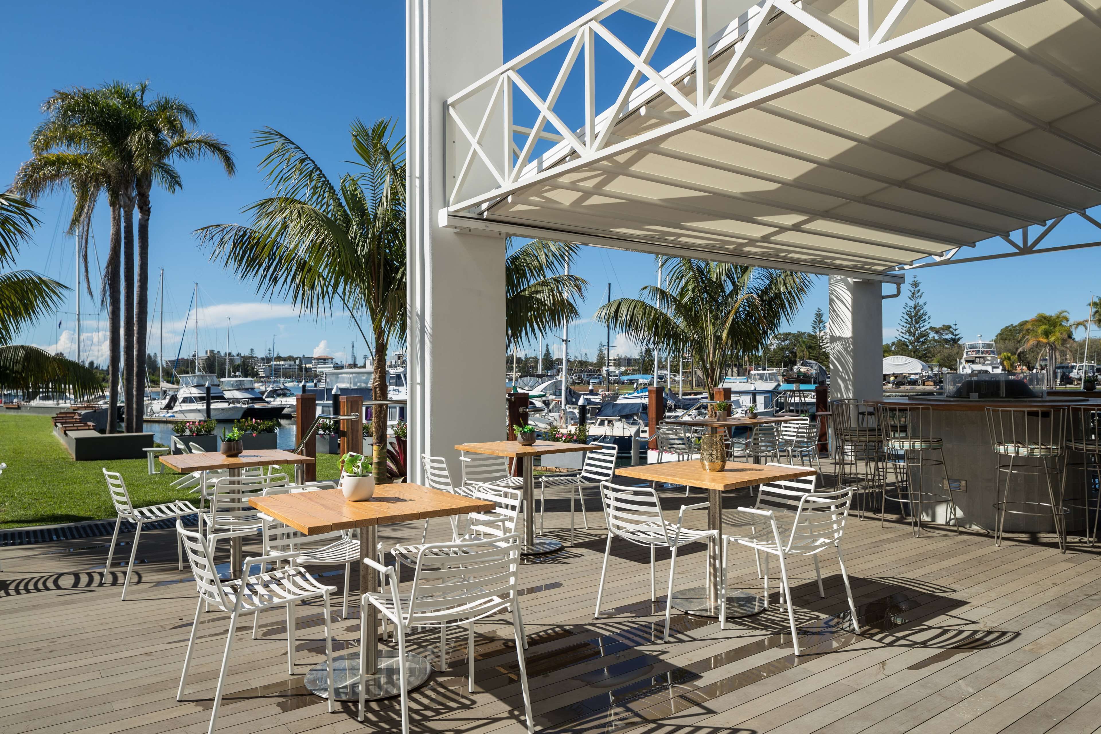 An outdoor patio with white metal chairs and tables, overlooking a marina with boats.