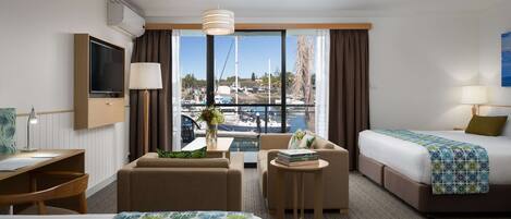 Ein Hotelzimmer mit zwei Betten, einem Schreibtisch, einem Stuhl, einem kleinen Tisch und Blick auf einen Hafen.