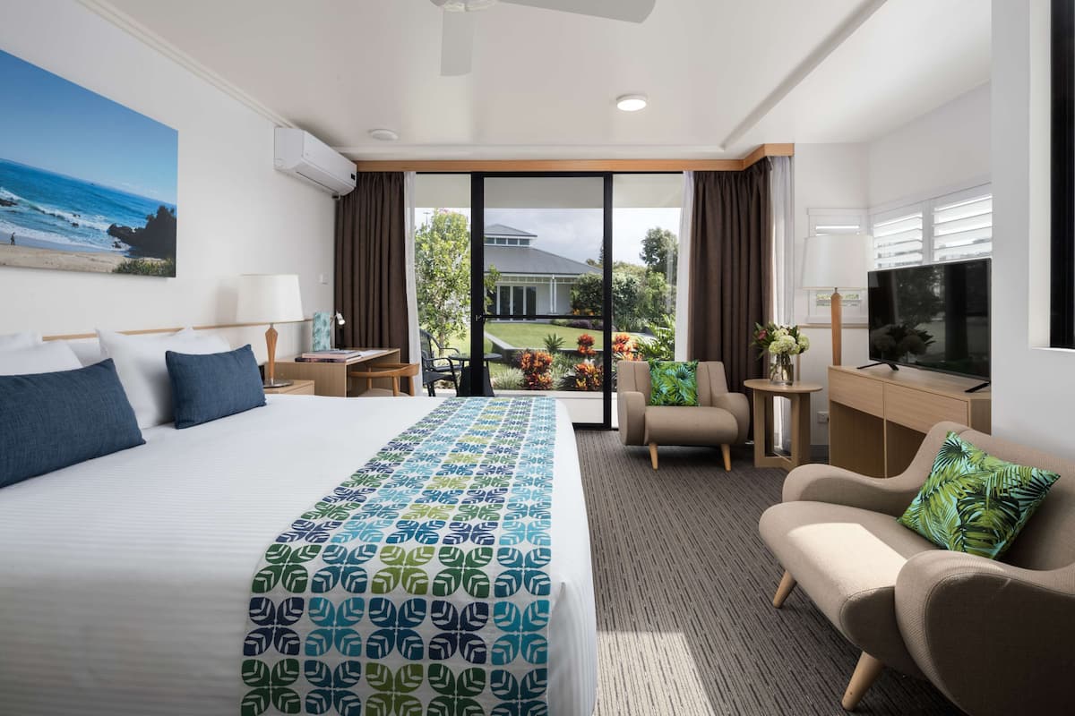 Une chambre d’hôtel avec un grand lit, un canapé, un bureau et un balcon offrant une vue sur l’océan et une maison.