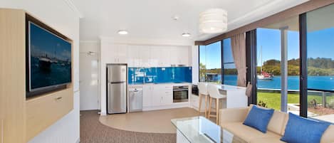 A modern kitchen with white cabinets, a stainless steel refrigerator, and a blue backsplash. There is a dining area with a glass table and beige sofa with blue cushions. The room has large windows offering a view of a lake and boats.