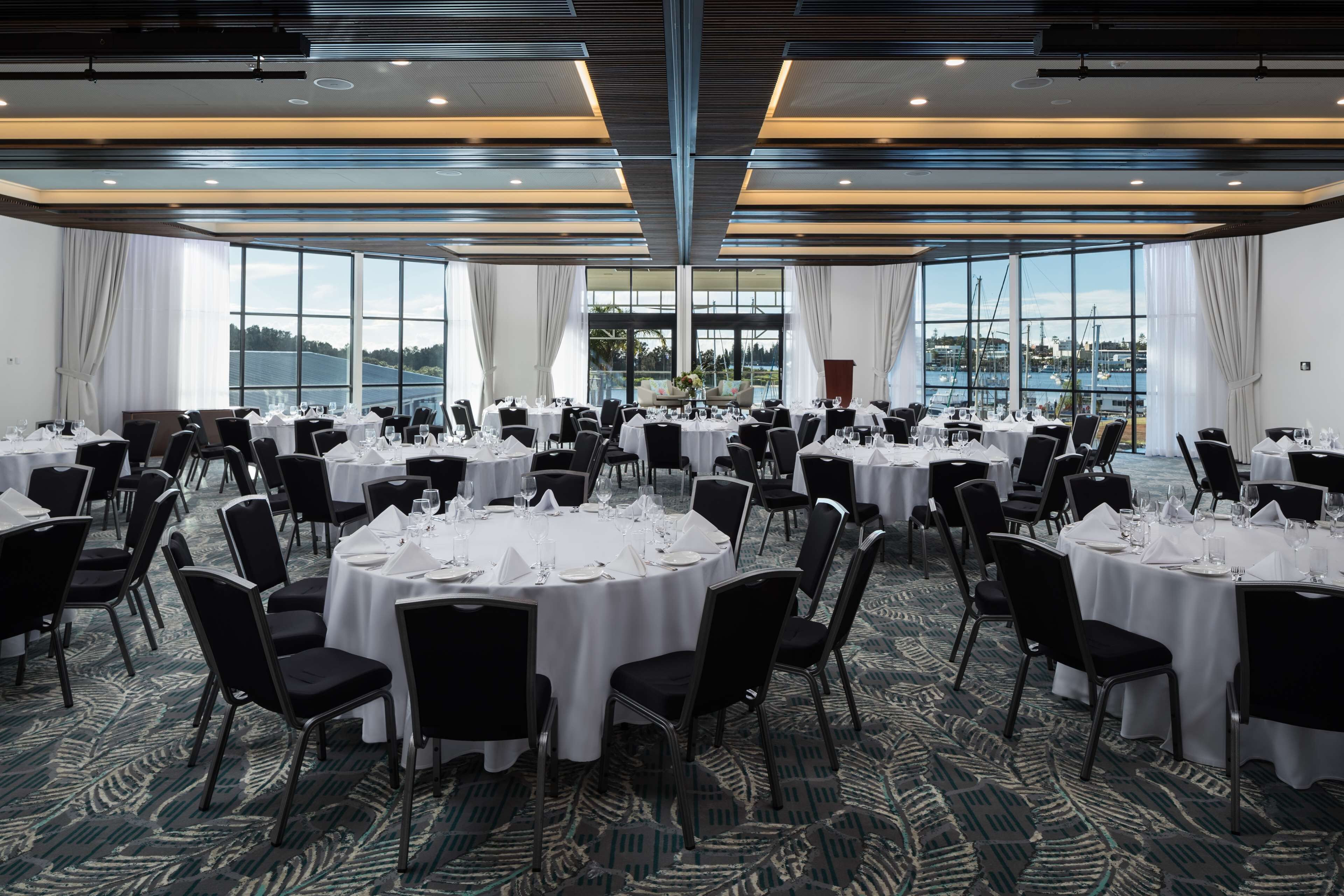 A large banquet hall with round tables set for dining, large windows offering a view of the sea, and a modern ceiling design.