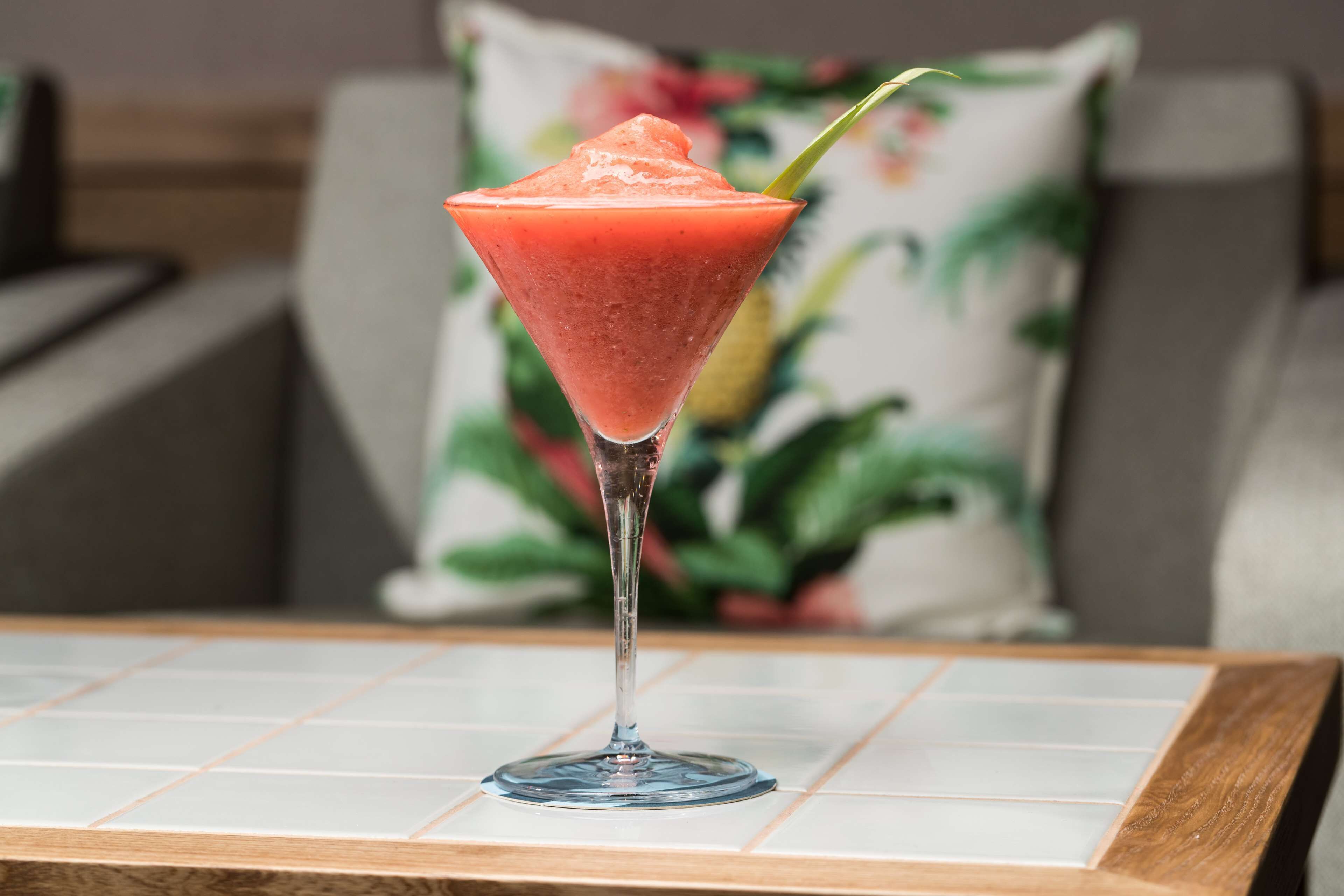 Un cóctel en una copa de martini sobre una mesa de baldosas, con un cojín floral al fondo.