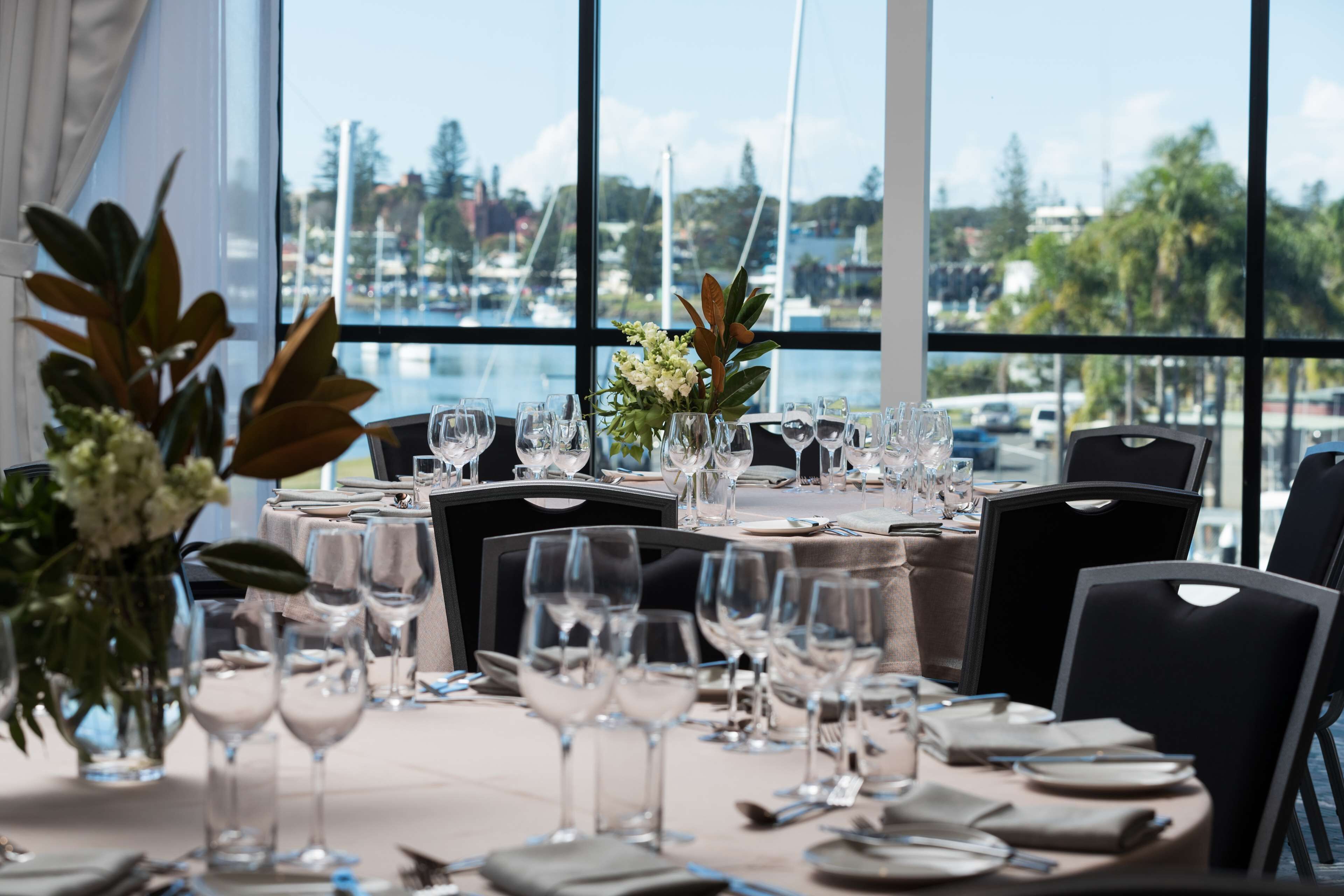 A dining table set with multiple wine glasses, plates, and cutlery, with a view of a marina and boats through large windows.