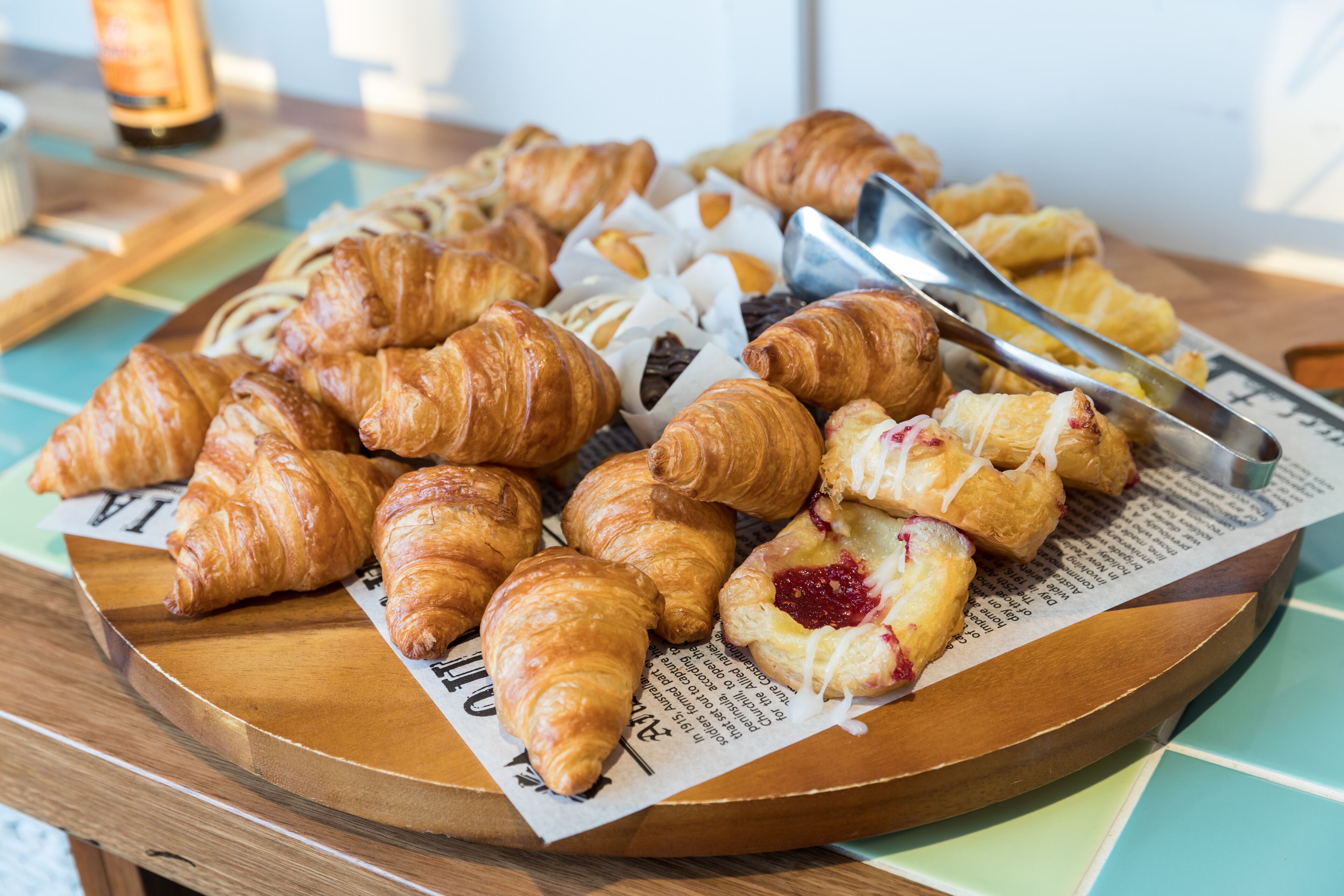 Variedad de pasteles en una bandeja de madera, incluyendo croissants y un pastel con relleno rojo, sobre un periódico.