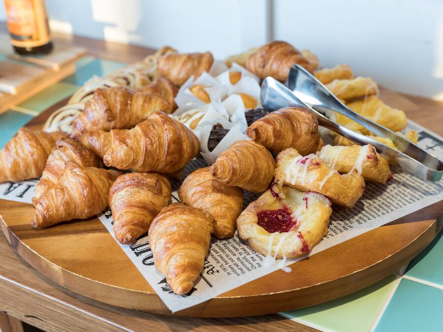 Variedad de pasteles en una bandeja de madera, incluyendo croissants y un pastel con relleno rojo, sobre un periódico.