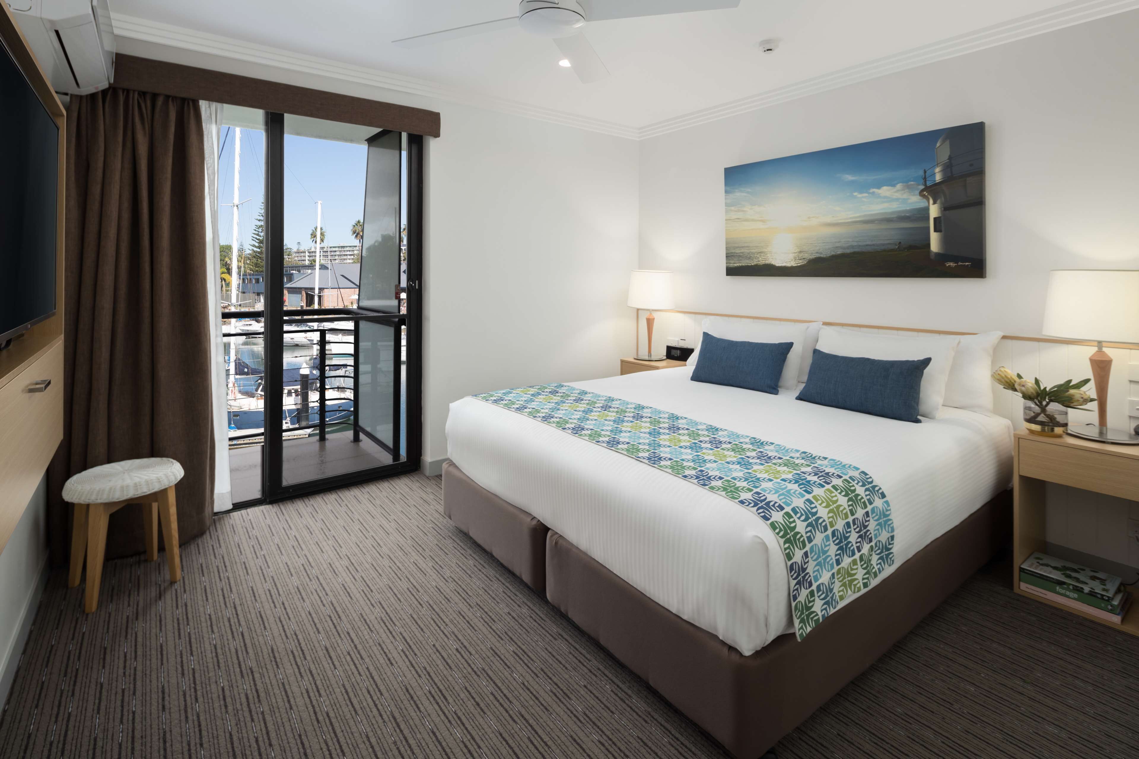 A hotel room with a large bed, a view of the outside, a painting on the wall, and a desk with a lamp.